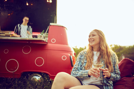 Focus on smiling, beautiful young woman sitting on floor pillow outdoors on picnic, drinking...
