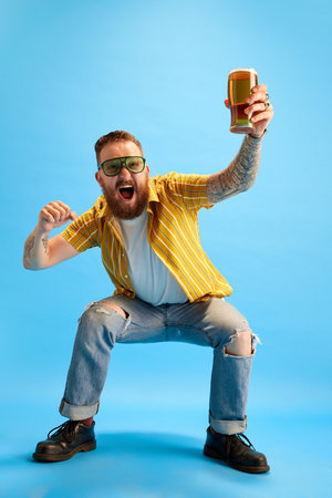 Portrait of emotional and excited bearded man in sunglasses and yellow shirt posing with beer against blue studio backgroundの写真素材