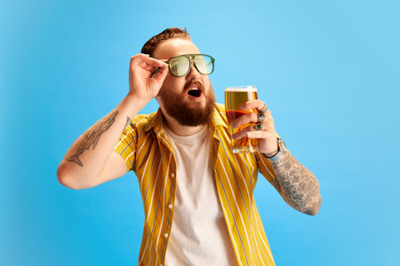 Bearded cheerful man in yellow shirt and sunglasses drinking beer against blue studio background....