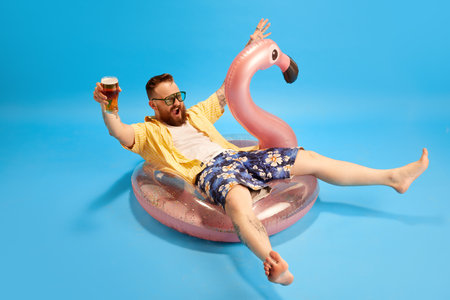 Happy and relaxed bearded man in swim shorts and yellow shirt sitting in swimming circle with beer...