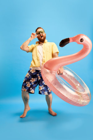 Happy and relaxed bearded man in swim shorts and yellow shirt standing with swimming circle against blue studio background. Summer vacationの写真素材