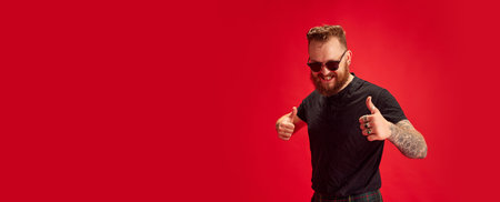 Portrait of bearded stylish man in sunglasses cheerfully posing against red studio background