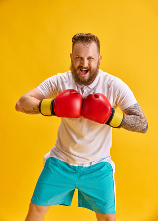 Bearded emotional man in sportswear and boxing gloves against bright yellow background. Active...