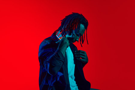 Musician. Handsome, stylish young guy with dreads posing against red background in neon lightの写真素材