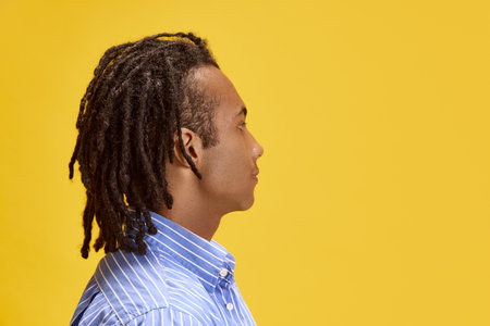Side view portrait of young african man with dreads in shirt looking straight against yellow studio...