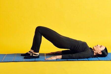Sportive woman in black sportswear lying on fitness mat and doing press against yellow studio...