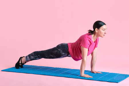 Slim mature woman training, standing in plank position, doing full body exercises against pink...