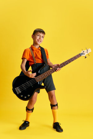 Boy, child in stylish clothes playing guitar against yellow studio background. Music and educationの写真素材