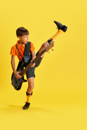 Emotional boy, child in stylish clothes playing guitar and dancing against yellow studio background. Music lifestyle and educationの写真素材