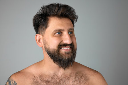 Bearded, smiling, mature, handsome man with blue eyes standing shirtless, looking away against grey studio backgroundの写真素材