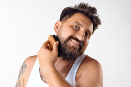 Mature, smiling, handsome man taking care after beard, brushing with special hairbrush against white studio backgroundの写真素材