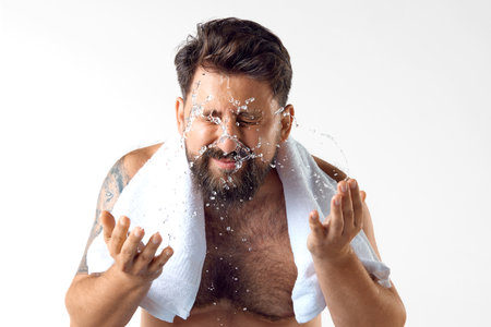 Morning get ready. Bearded shirtless man with towel on shoulders, washing face against white studio backgroundの写真素材