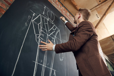 Bottom view of young man, fashion designer creating new collection of clothes, drawing suit sketches on blackboardの写真素材