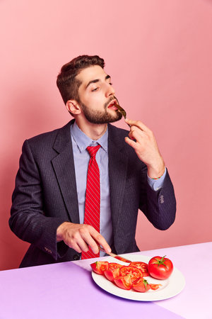 Businessman eating tomatoes with knife and fork against pink background. Vegetables, vitamins, freshnessの写真素材