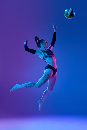 Full-length of young sportive woman, volleyball player in motion, hitting ball in a jump against blue studio background in neon lightの写真素材