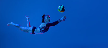 Sportive young woman, volleyball player in motion, hitting ball, competing against blue studio background in neon lightの写真素材