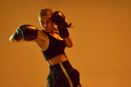 Sportive teen girl, MMA athlete in uniform and boxing gloves, training against orange studio background in neon lightsの写真素材