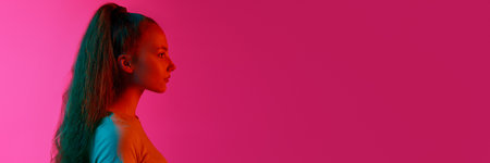 Profile view of young girl looking straight with calm emotionless face against pink studio background in neon lightの写真素材