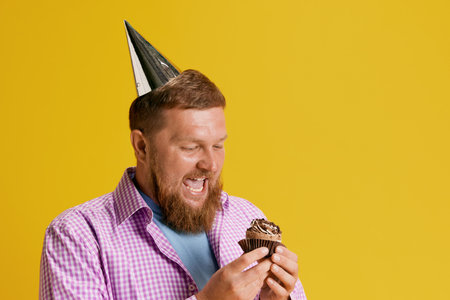 Excited bearded man feeling happy and positive, eating his birthday cupcake against yellow studio backgroundの写真素材