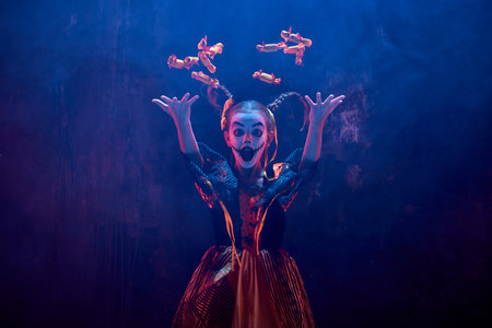 Little girl, child in costume and makeup for halloween throwing sweets away against dark background...