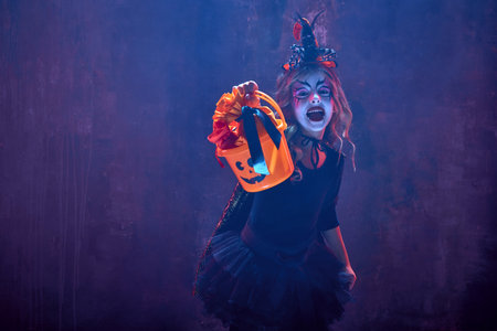 Little girl, child in image of witch, for halloween party standing with basket for sweets against...