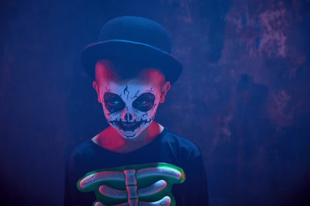 Little boy, child with creepy pale face makeup, wearing costume of skeleton against dark background in neon light with smokeの写真素材