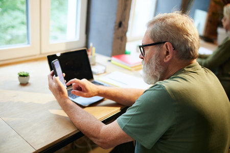 Senior businessman sitting at table in office, scrolling mobile phone screen, working with online servicesの写真素材