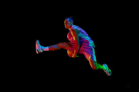 Competitive teen boy in motion, playing, training basketball, jumping with ball against black studio...