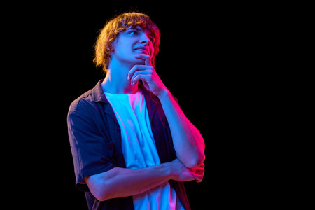 Portrait of young guy, student in casual clothes posing with thinking face against black studio background in neon lightsの写真素材