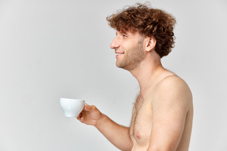 Morning coffee. Side view image of smiling, shirtless, mature man with curly hair standing with cup against grey studio backgroundの写真素材