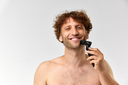 Portrait of smiling, mature man with curly hair, using razor for shaving against grey studio...