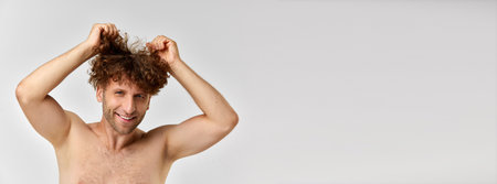 Messy hair. Positive mature man standing shirtless and taking care after curly hair against grey studio backgroundの写真素材
