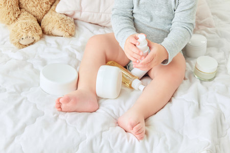 Cropped image of child legs, toddler sitting in bed with body moisturizing creams, lotions. Skincare productsの写真素材