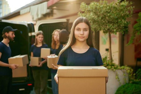 Focus on girl, volunteer standing outdoors near van on sunny day. Young people on background loading boxes into vanの写真素材
