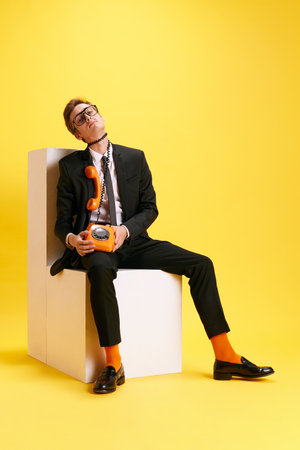 Young stylish guy. businessman in black suit sitting with telephone around him against yellow studio background. Deadlinesの写真素材