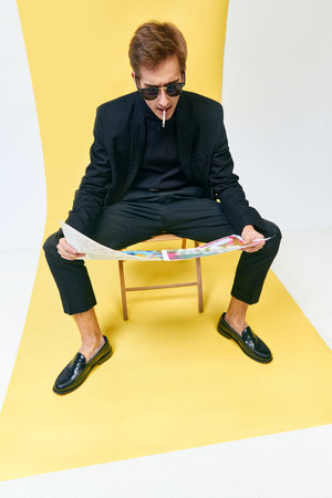 Handsome stylish man in black suit and sunglasses, writer, journalist reading newspaper against yellow studio backgroundの写真素材
