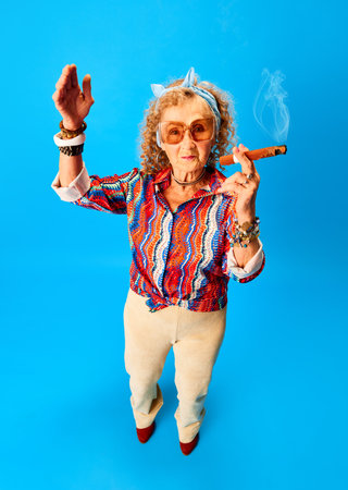 Cheerful, positive old woman in stylish clothes and sunglasses smoking cigar over blue studio...