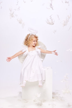 Happy, smiling, positive little girl, child in costume of angel sitting isolated over white studio background with flying feathersの写真素材