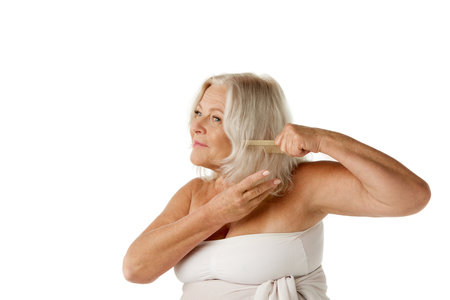 Beautiful senior woman in her 60s taking care after hair, standing in shower towel and brushing hair against white studio backgroundの写真素材