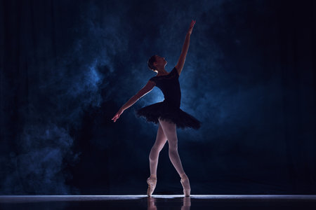 Beautiful, elegant young woman, professional ballet dancer in tutu dancing on stage over dark...