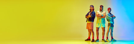 Three stylish young men in colorful vintage sportswear standing against gradient yellow blue background in neon lightの写真素材