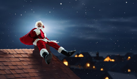 Cheerful, happy senior man Santa Claus sitting on chimney on roof at night with big bag with presentsの写真素材