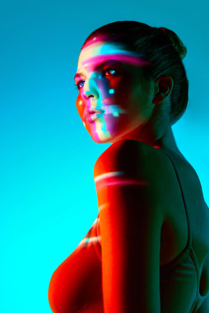 Portrait of beautiful young woman against blue background with multicolored neon reflection on face....