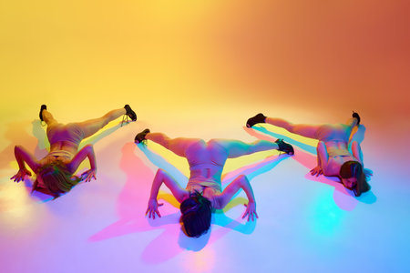 Freedom of movements. Three young woman in underwear dancing high heel dance on floor over gradient yellow background in neon lightの写真素材