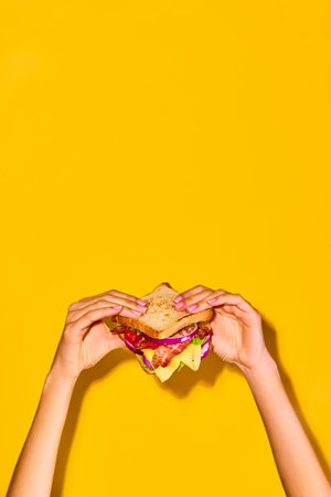 Woman holding delicious sandwich with grilled bread, ham, lettuce, cheese, tomato, onion and avocado over yellow background. Cooking, taste. Posterの写真素材