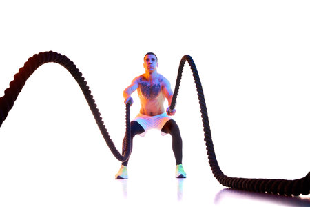 Shirtless, sportive, muscular young man training with battle rope against white studio background in...