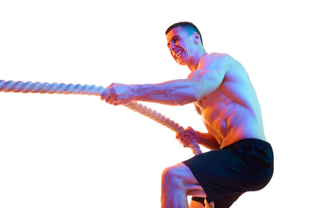 Shirtless young man with strong body and hands pulling rope, training against white studio...