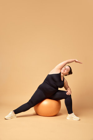 Stretching and balance. Young overweigh woman in black sportswear training with fitness ball against beige studio background. Sport, body-positivityの写真素材
