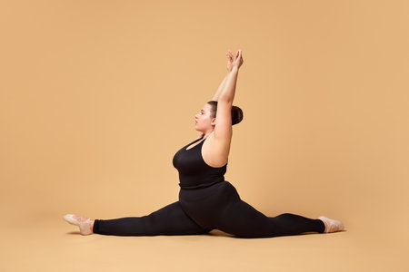 Young overweight woman in black sportswear training, sitting on twine against beige studio background. Sport, body-positivityの写真素材