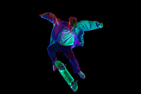 Teen boy, active guy in casual clothes in motion, training, practicing stunts on skateboard against black studio background in neon lightの写真素材